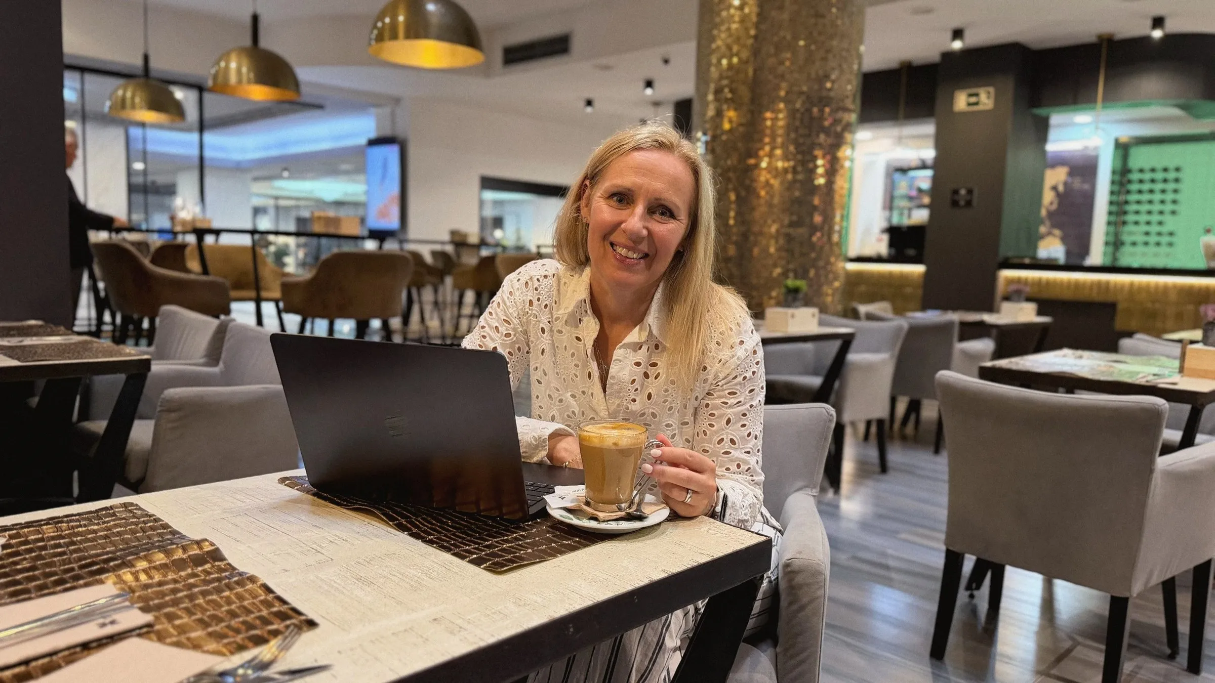 Kirsten smiling at a café table with a coffee and her laptop