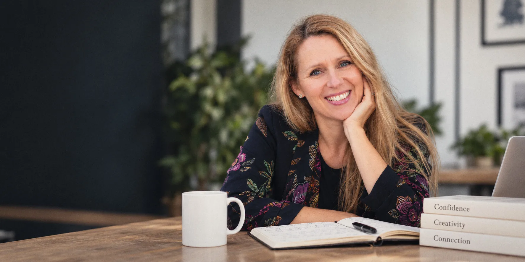 Kirsten smiling at a table with a notebook, a mug and three books: Confidence, Creativity, Connection